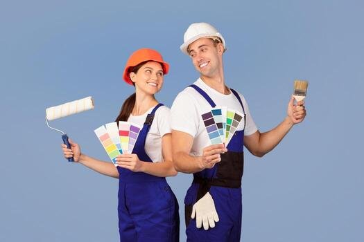 cheerful-couple-in-hard-hats-posing-with-paint-brushes-and-color-swatches-over-blue-studio-background-young-family-in-work-clothes-choosing-palette-to-repair-their-home-ready-for-renovation-photo